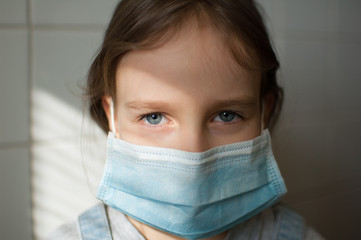 Little sad girl in grey T-shirt wearing a protective disposable mask stay home because of the Coronavirus Covid-19 in self-isolation on white tiles background on the kitchen