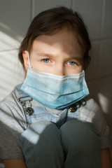 Little sad girl is restricted at home because of the pandemia of Coronavirus Covid-19 and self-isolation. Kid is sitting at the kitchen on white tiles background in a protective disposable mask