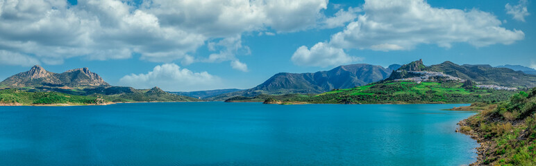 View of Zahara de la Sierra a white town or Pueblos Blancos, in Andalucia Spain, with a ruined medieval castle. Man made turquoise water lake formed by the Guadalete river high damn in foreground