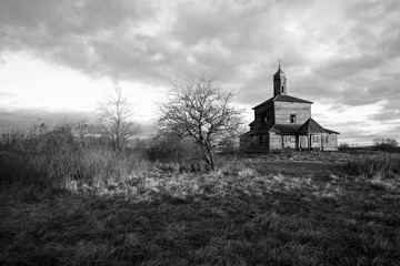 Black and white picture of an old abandoned wooden church in the field behind the trees. Cloudy sky at the background