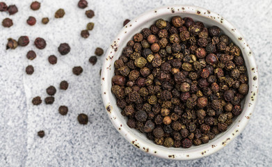 Black Organic dried Peppercorns in a white bowl on a light concrete or stone background close-up. Spices and seasonings. Selective focus, top view and copy space