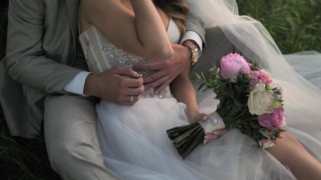 Wedding couple. The groom hugges the bride. Hugging newlyweds sitting on the grass. Bride and groom embracing outdoors. Man and woman in love. Bride holding her wedding bouquet of peonies and roses.