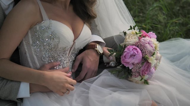 Wedding couple. The groom hugges the bride. Hugging newlyweds sitting on the grass. The groom holds the bride's bouquet of peonies and roses. Bride and groom embracing outdoors. 