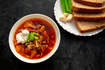 Borscht soup in a white bowl with black bread, green onion and garlic on dark gray background. Famous traditional Ukrainian and Russian borshch soup with beetroot and cabbage. Red Eastern Europe soup.