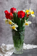 Bouquet of tulips and daffodils in a crystal vase on a concrete wall background.