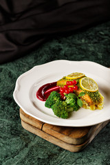 Healthy air fry salmon sprinkled with steamed broccoli, cranberry and sliced lemon in a white plating. Food photography in dark background for cook book concept.