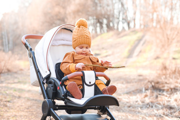Baby boy in warm jacket and hat sitting in modern stroller on a walk in a park.