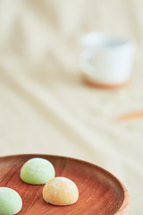 Three bright colorful mochis on a wooden plate and a coffee cup on a beige fabric.