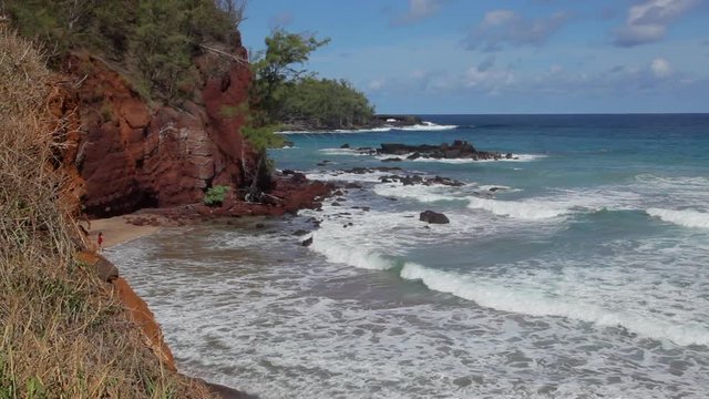 Red cliffs of Koki Bay past Hana on the drive of the famous Hana Highway on Maui showing waves rolling to the lava-rock shore with lava outcroppings in the ocean on a sunny blue-sky Hawaii day.