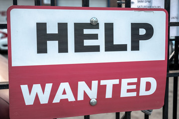 A red black and white sign with help wanted text hanging on a black cast iron fence along the sidewalk with two bolts in Chicago during the COVID-19 outbreak and pandemic.