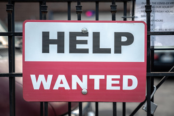 A red black and white sign with help wanted text hanging on a black cast iron fence along the sidewalk with two bolts in Chicago during the COVID-19 outbreak and pandemic.