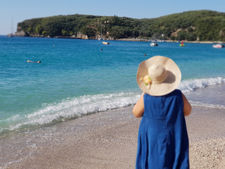 Fototapeta na wymiar Back view of the young woman in straw hat and blue dress relaxing on the beach and looking forward into the lagoon in Parga Greece.