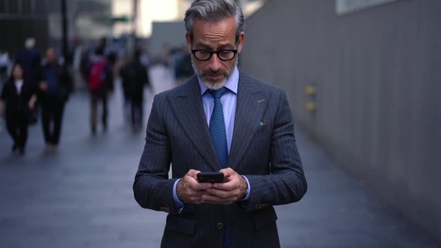 Confident businessman in formal wear chatting online on website on smartphone while strolling to office, successful executive director 50 years old reading incoming notification on cellular phone
