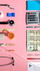 Medical and Financial Supplies face off on light pink surface. Money, insulin, pills, bandage, calculator, pen, test tube, stapes, stethoscope.