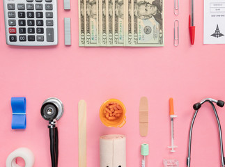 Medical and Financial Supplies face off on light pink surface. Money, insulin, pills, bandage, calculator, pen, test tube paper clips, syringe.