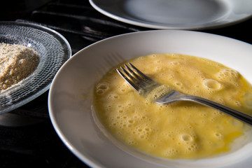 Raw scrambled eggs on a white plate and a fork, ingredients to prepare breaded chicken, fish, shrimp, beef in a home kitchen, culinary concepts and recipes.