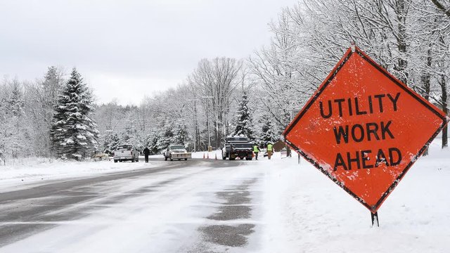 UTILITY WORK AHEAD sign warns traffic about a work crew on a snowy city street, on a frosty winter day. Frost and snow covered trees and pickup truck with blade are seen.