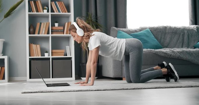 Active young girl in sport clothing repeating exercises for legs while watching workout tutorial on laptop. Concept of active and healthy lifestyle.