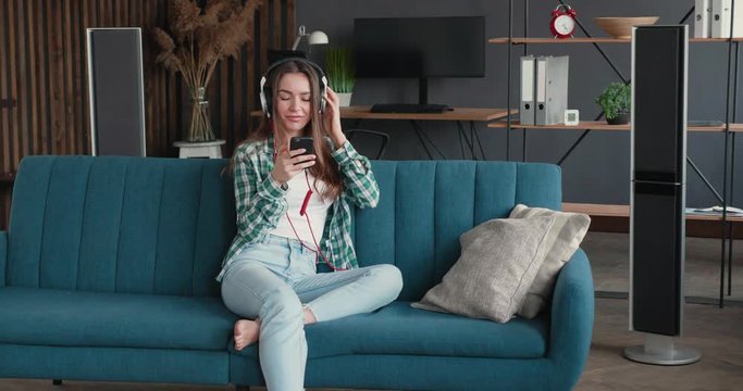 Attractive Woman is turning on her Favourite Music and started to relax on the Sofa. Young casual Woman is listening to Music in big Headphones, using Musically Apps for Smartphones. Music. Good Mood.