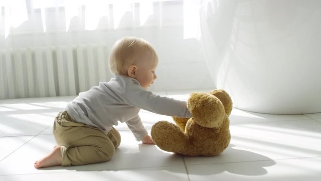 Handheld tracking shot of adorable baby girl with blond hair crawling on floor and taking plush toy bear