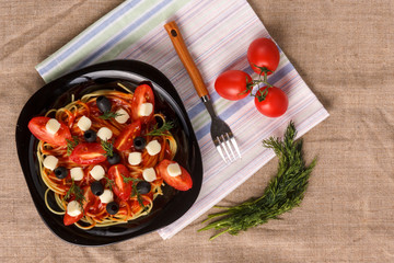 Black plate with spaghetti in a sauce with cherry tomatoes olives and mozzarella on a napkin on a linen tablecloth.