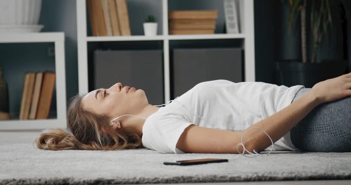 Tired young girl on white t-shirt and grey leggings relaxing on floor in earphones after morning exercise. Sporty woman with blond hair taking break after workout at home.
