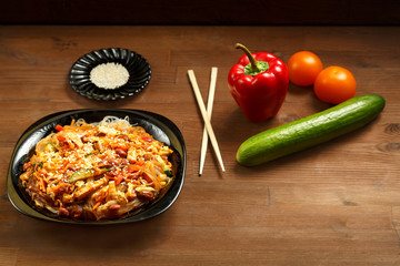 Black plate with funchose and chicken with vegetables under pad Thai sauce on a wooden table and fresh vegetables.