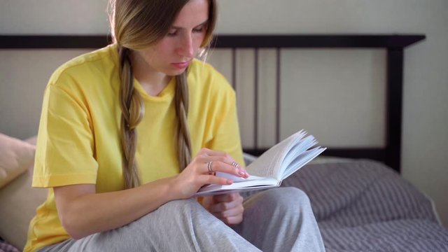 A girl with pigtails in a yellow t-shirt and home clothes is sitting on the bed, reading a book, thinking and dreaming. Close-up 4K