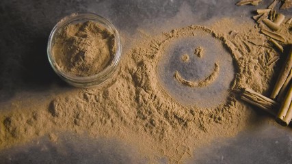 A fun happy face drawn in powdered cinnamon on a countertop along with fresh cinnamon sticks as a breeze blows the design away.