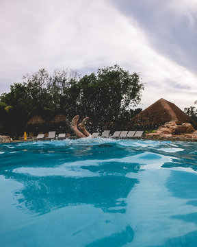 Feet of man at beautiful pool with buildings with straw roof, trees, and cloudy sky, Mexico