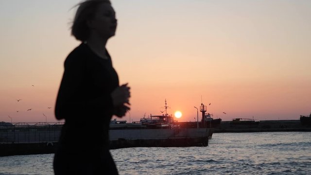 The girl daily runs evening jogs along the promenade against the backdrop of the sea pier and sunset. The wind accelerates the waves. Seagulls fly over the waves.