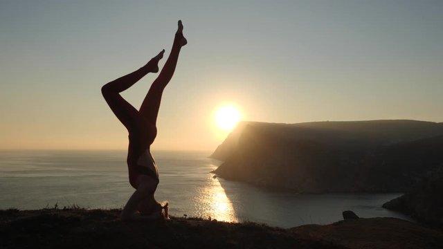 The girl practices yoga. Headstand on the background of the sea and mountains. Sunset over the coastal hills.
