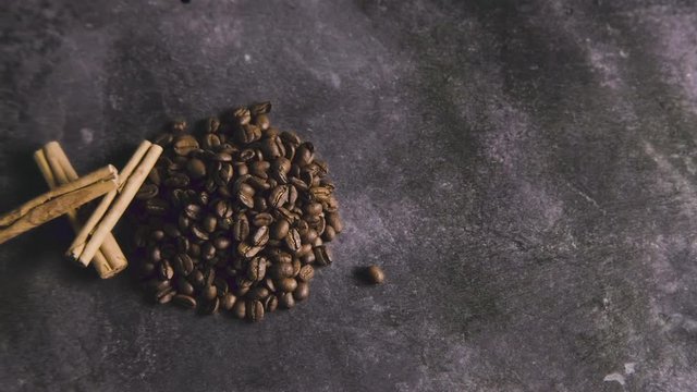 Top-down pan over a small pile of coffee beans with several sticks of cinnamon on a grey granite background.