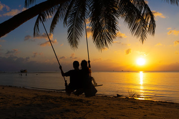 Fototapeta na wymiar Silhouetted couple in love walks on the beach during sunset. Riding on a swing tied to a palm tree and watching the sun go down into the ocean