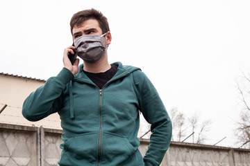 A man in a gray medical mask outdoors. He is talking on the phone.
 He is dressed in a green sweater. In the background is a gray, concrete fence, grass and trees. Quarantine time.