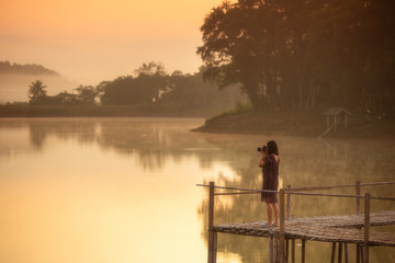 Asia woman photographer take photo on bamboo bridge in morning with orange light of sunset.