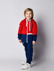 Full growth portrait of happy smiling blond kid boy in red and blue sportwear and white sneakers