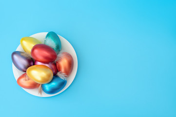 Decorated Easter eggs with small feathers lie on the plate on blue background. Happy Easter holiday concept. Greeting, invitation card. Flat lay style with copy space.