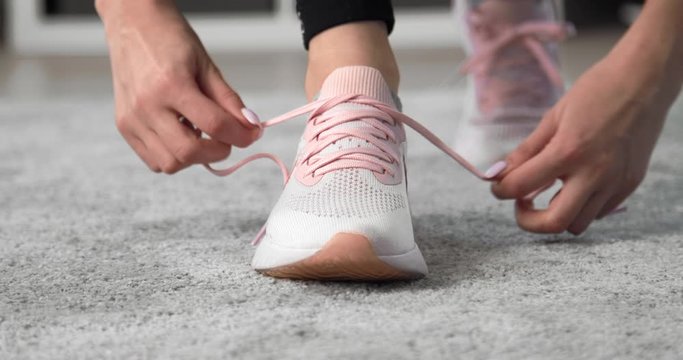 Close up of fitness girl tying laces on sport sneakers on grey carpet. Young woman preparing for training at home. Focus on shoes.