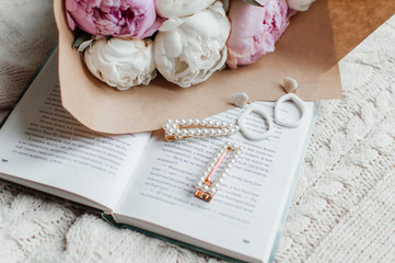 A beautiful bouquet of white and pink peonies lie on an open book. On the book next to the bouquet are beautiful white earrings and hair clips. Vintage hair clips and earrings. Present.
