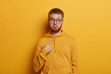 Indignant serious hipster indicates at himself with wondered expression, asks who me, wears optical glasses and sweatshirt, isolated on yellow background. Puzzled guy heard someone mentioned his name