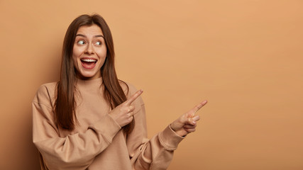 Photo of cheerful adult woman sells product, points at empty space aside, makes choice and smiles pleased, wears casual jumper, suggests pick item in store, gestures against brown background