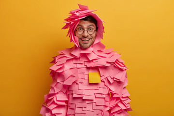 Photo of happy young European man with pleasant smile, cheerful expression, glad to finish work in time, receives praise, stands covered by sticky notes, stands indoor against yellow background