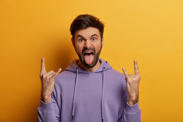 Crazy bearded hipster enjoys rock party, makes heavy metal sign, enjoys cool music, exclaims, brings positive vibes, dressed in purple sweatshirt, isolated on yellow background, sticks out tongue