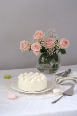 White cream cake on the table with a white tablecloth. With a bunch of pink roses and a stack of white plates