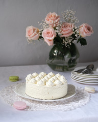 White cream cake on the table with a white tablecloth. With a bunch of pink roses and a stack of white plates
