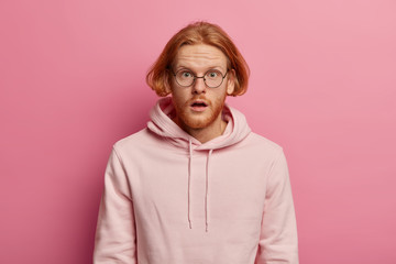 Surprised excited bearded foxy guy feels astonished, reacts on unbelievable news, stares through transparent glasses and dressed in sweatshirt poses against rosy pastel background being very impressed