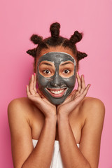 Young pleased woman applies clay mask for beauty, touches cheeks gently, enjoys softness of skin, stands wrapped in towel after taking bath, smiles pleasantly, has anti wrinkle procedures at home