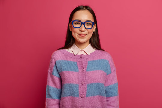Pretty young woman student looks at camera determined and happy, wears transparent glasses and striped jumper, feels proud to get excellent mark, receives praise, isolated on pink background