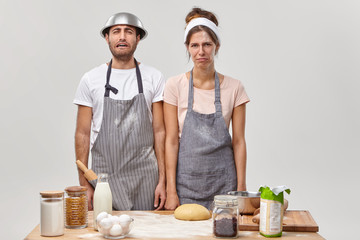 Tired discontent housewife and husband stand together at kitchen near table with ingredients, make dinner for family, prepare dough for baking pizza, exhausted of routine activity, wear aprons